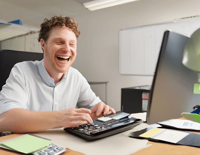 Budgeting Tips for Responsible Gambling on Online Slot Websites A white man sitting at a desk smiling and focused on his laptop screen. Next to it a calculator rests its buttons catching the overhead light in sharp relief - Budgeting Tips for Responsible Gambling on Online Slot Websites